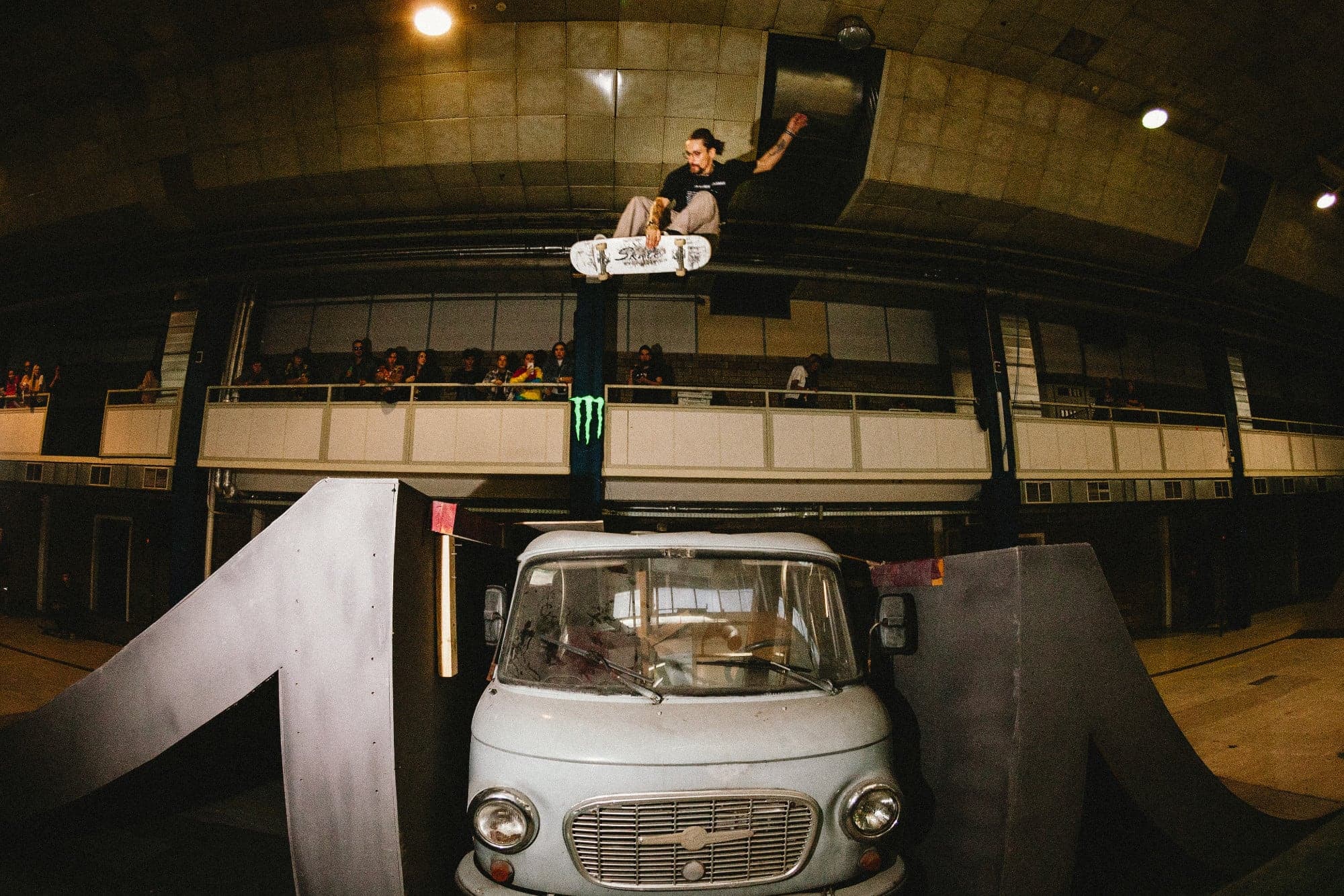 Skateboarder airing over a van built into the course at an indoor contest while the crowd watches from the balcony