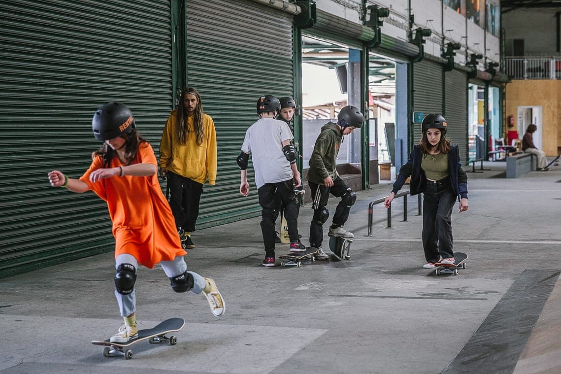 Group of students rolling across the flat of a covered skatepark while their coach watches